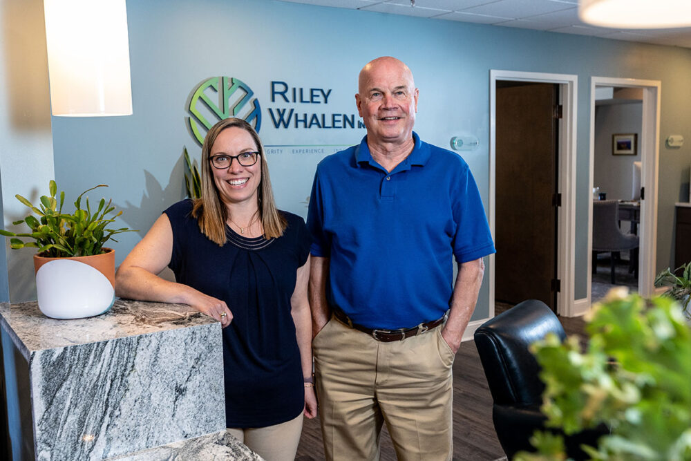 a man and a woman standing in an office with the logo of their business "Riley Whalen" on the wall behind them.