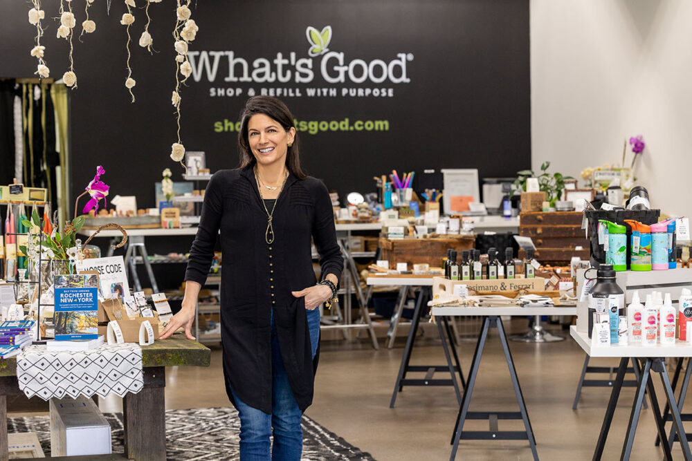 A woman standing with her hand on a table in an eclectic store with the logo for the business "What's Good" on the wall behind her.